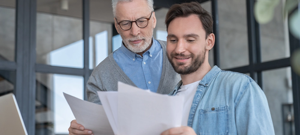 Caring loving adult son helping his old senior elderly father paying bills, counting money, doing paperwork at home together. Aid to elderly generation