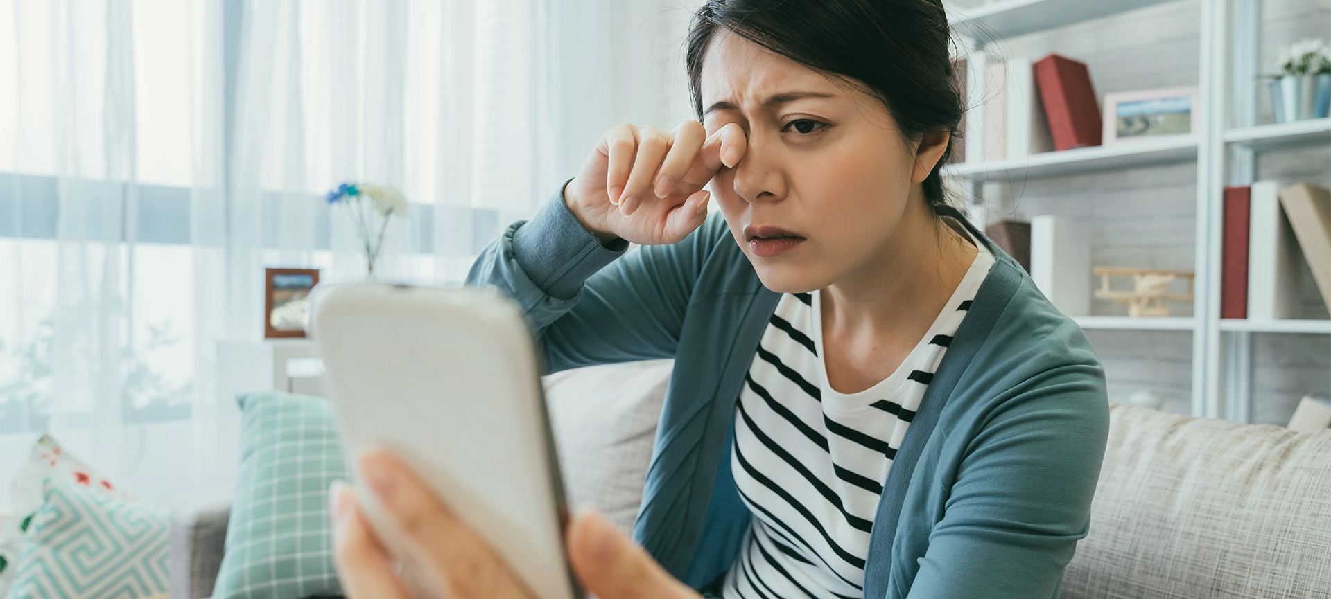 selective focus asian female suffering blurred vision is rubbing her tired eyes while holding using her smartphone on the couch in the living room at home.