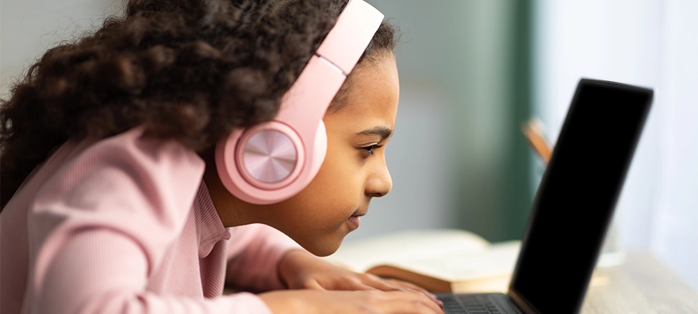 Youth and technology concept. Focused black schoolgirl using laptop computer, sitting at table too close to pc, side view. Little girl with poor eyesight having near vision problem