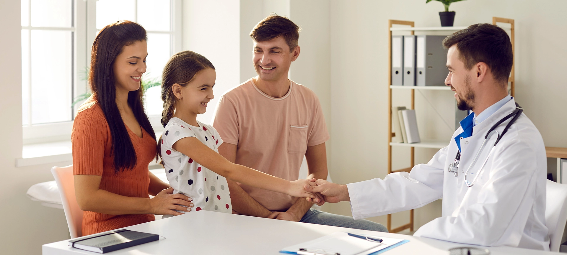 Smiling little girl shaking hand to positive man doctor pediatrician after successful checkup in medical clinic with parents sitting around. Medicare for kids and family doctor concept