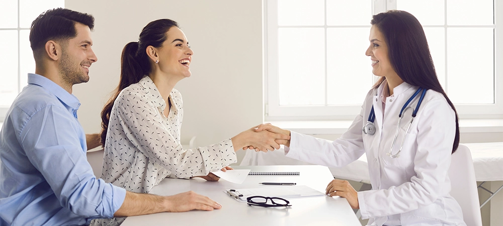 Happy woman patient giving gratitude handshake to doctor get good news about pregnancy on appointment at clinic office. Satisfied caucasian family couple visit physician for healthcare consultation