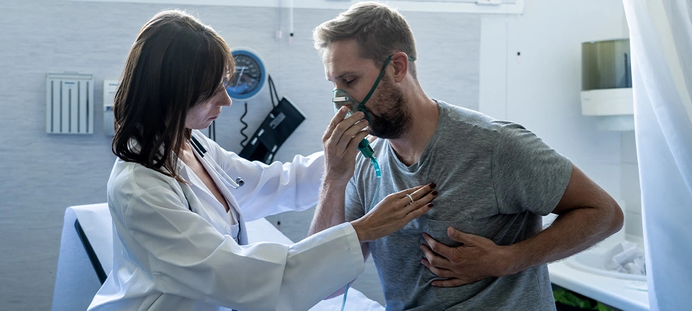 Young sick man patient with Oxygen Mask while female doctor listens his chest with stethoscope in hospital emergency room. In Smoking and respiratory diseases and anti tobacco advertising campaign.