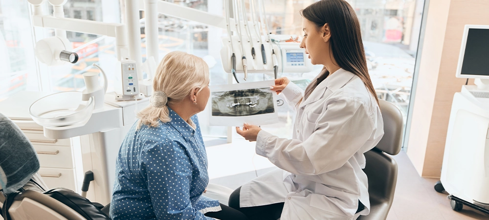 Elderly woman in blue polka-dot shirt listening to female doctor explaining dental X-ray, seated in dental office with equipment and tools. Concept of healthcare and pensioners, medicine.