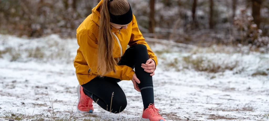 Close-up of a female runner clutching her knee, having stopped on a snowy trail. Sports injury or sudden training setback in cold winter weather.