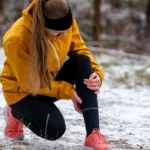 Close-up of a female runner clutching her knee, having stopped on a snowy trail. Sports injury or sudden training setback in cold winter weather.