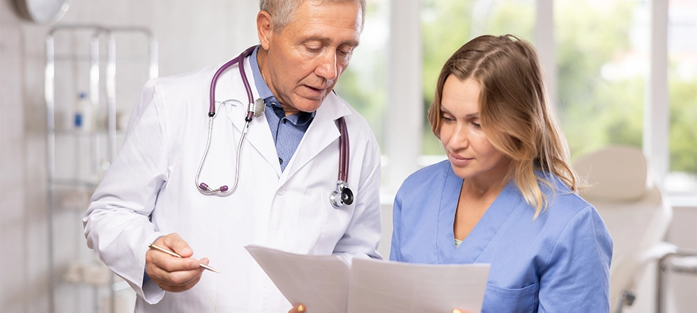 Two fellow doctors, adult woman and senior man, standing in medical office with papers in hands, focused on discussing of clinical diagnosis of patient
