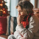 Young teen boy with long hair thoughtful look sad eyes negative mood angry and crying at home. Stylish zoomer gen Z pensive on new year holidays with xmas tree bokeh lights garlands eve 25 december