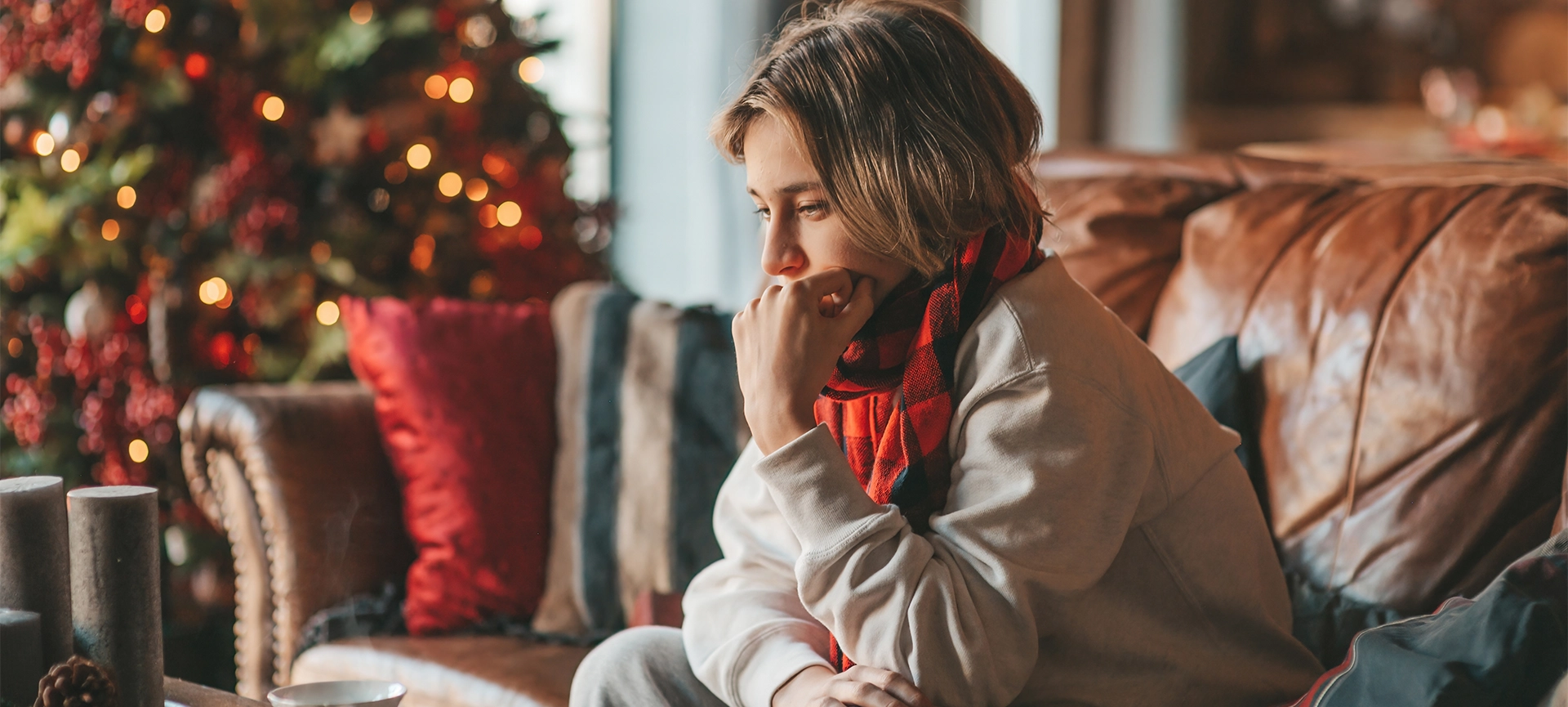 Young teen boy with long hair thoughtful look sad eyes negative mood angry and crying at home. Stylish zoomer gen Z pensive on new year holidays with xmas tree bokeh lights garlands eve 25 december
