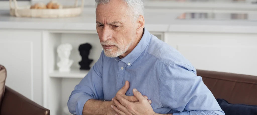 Concept of health and cardiovascular problems. Senior man with cardiology disease feeling sharp heart pain, holding hands on chest, sitting on armchair at home. Pensioner need emergency medical care