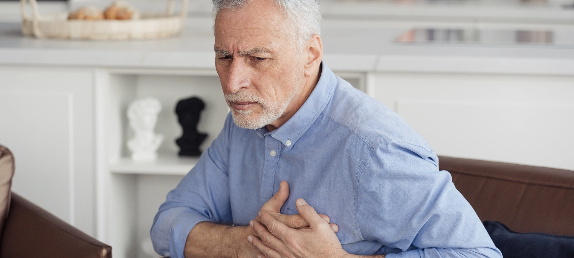 Concept of health and cardiovascular problems. Senior man with cardiology disease feeling sharp heart pain, holding hands on chest, sitting on armchair at home. Pensioner need emergency medical care