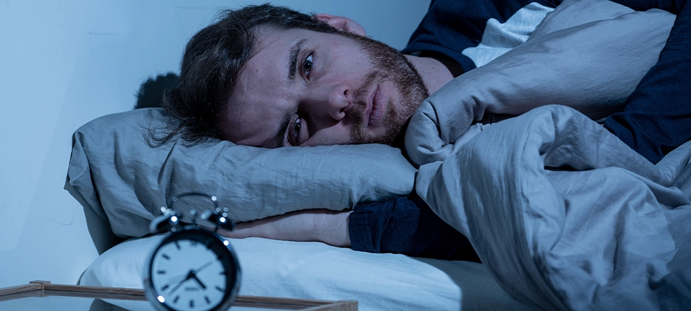 Sleepless and desperate young caucasian man awake at night not able to sleep, feeling frustrated and worried looking at clock suffering from insomnia in stress and sleeping disorder concept.