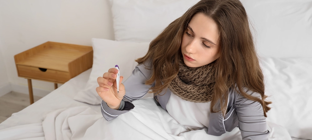 Ill young woman with thermometer sitting in bedroom