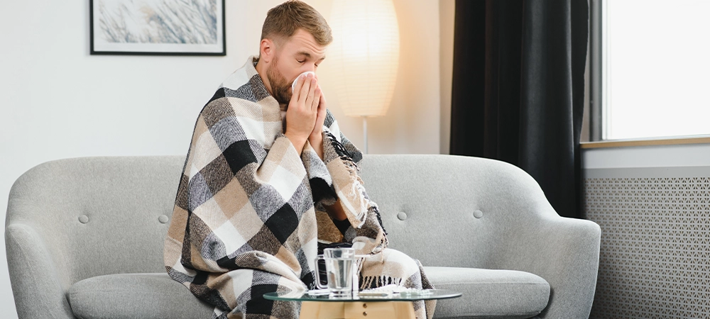 Sick adult man who has bad cold or seasonal flu virus is fighting illness at home. Unhappy middle aged man is sitting on sofa wrapped in warm scarf and plaid, holding paper tissue and sneezing